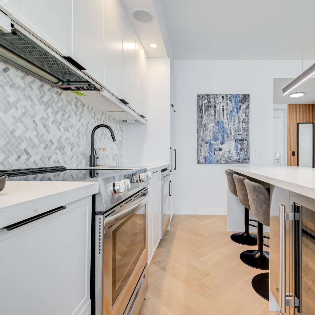 A modern kitchen featuring stainless steel appliances, white cabinetry, and a large countertop with four bar stools nearby.