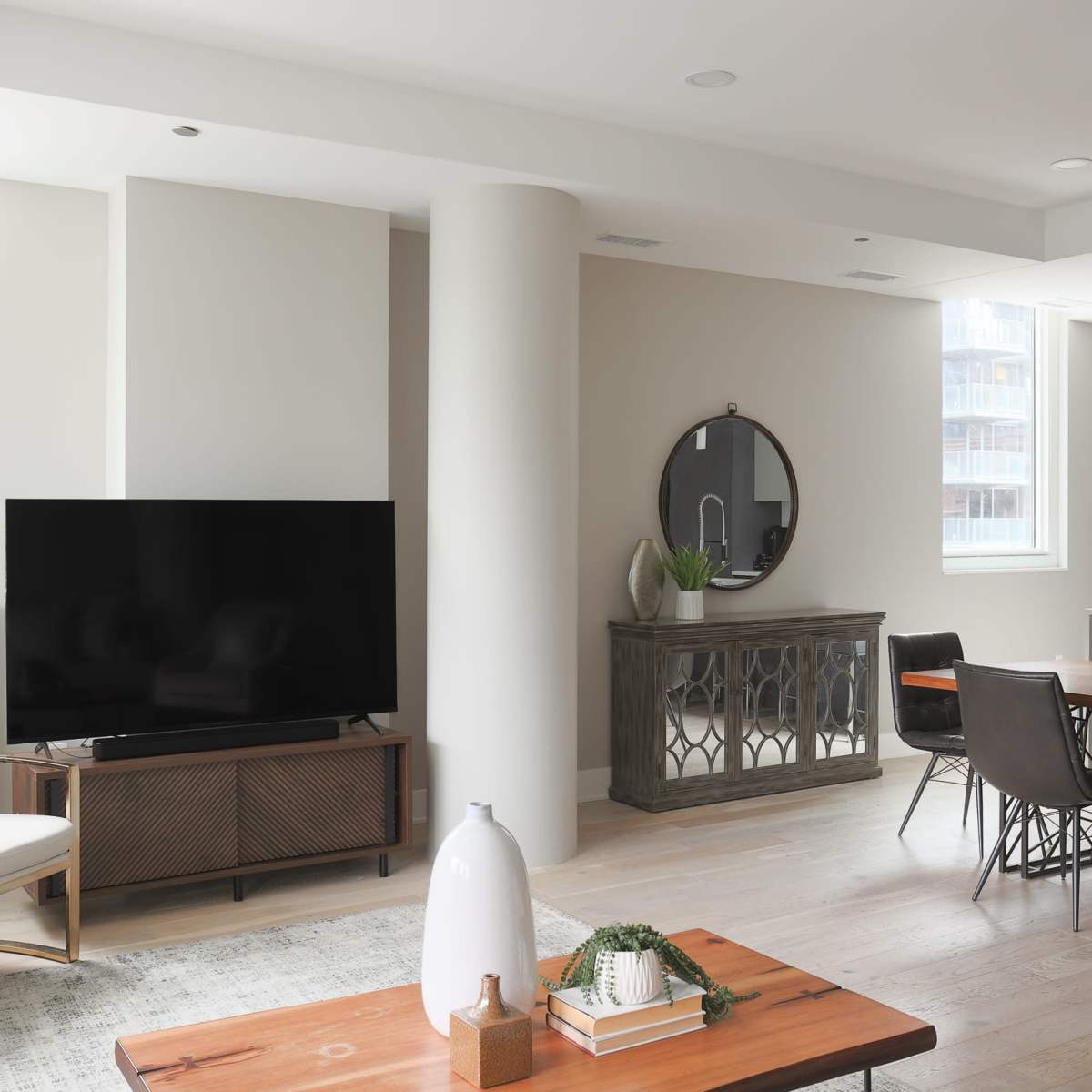 A modern living room featuring a television on a wooden media console, a round mirror on the wall, a dining area with dark chairs, and a coffee table with decorative items.