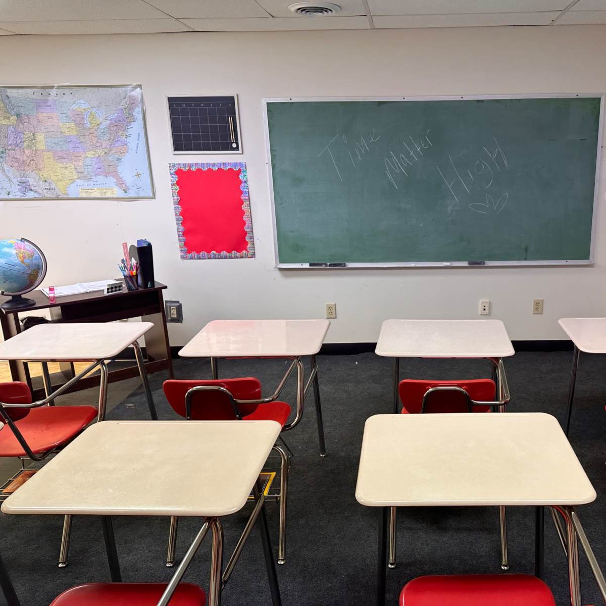 A classroom is arranged with several empty desks facing a chalkboard, surrounded by maps on the walls.