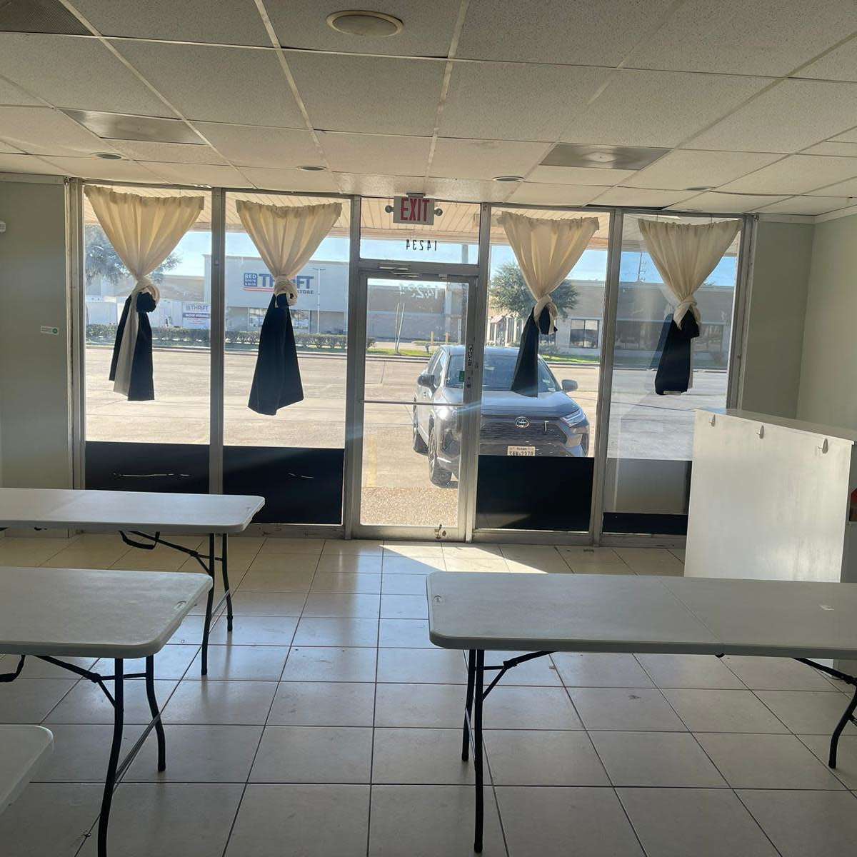 The interior of a vacant commercial space features several folding tables arranged in the center, large windows with curtains, and a view of a parked car outside.