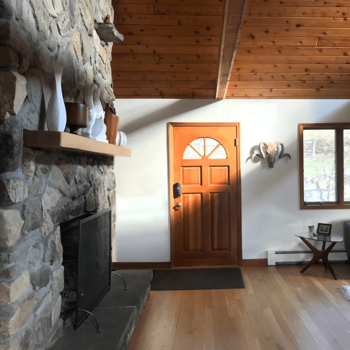 A stone fireplace is positioned next to a wooden door, with a large window and a gray sofa in a sunlit living room.
