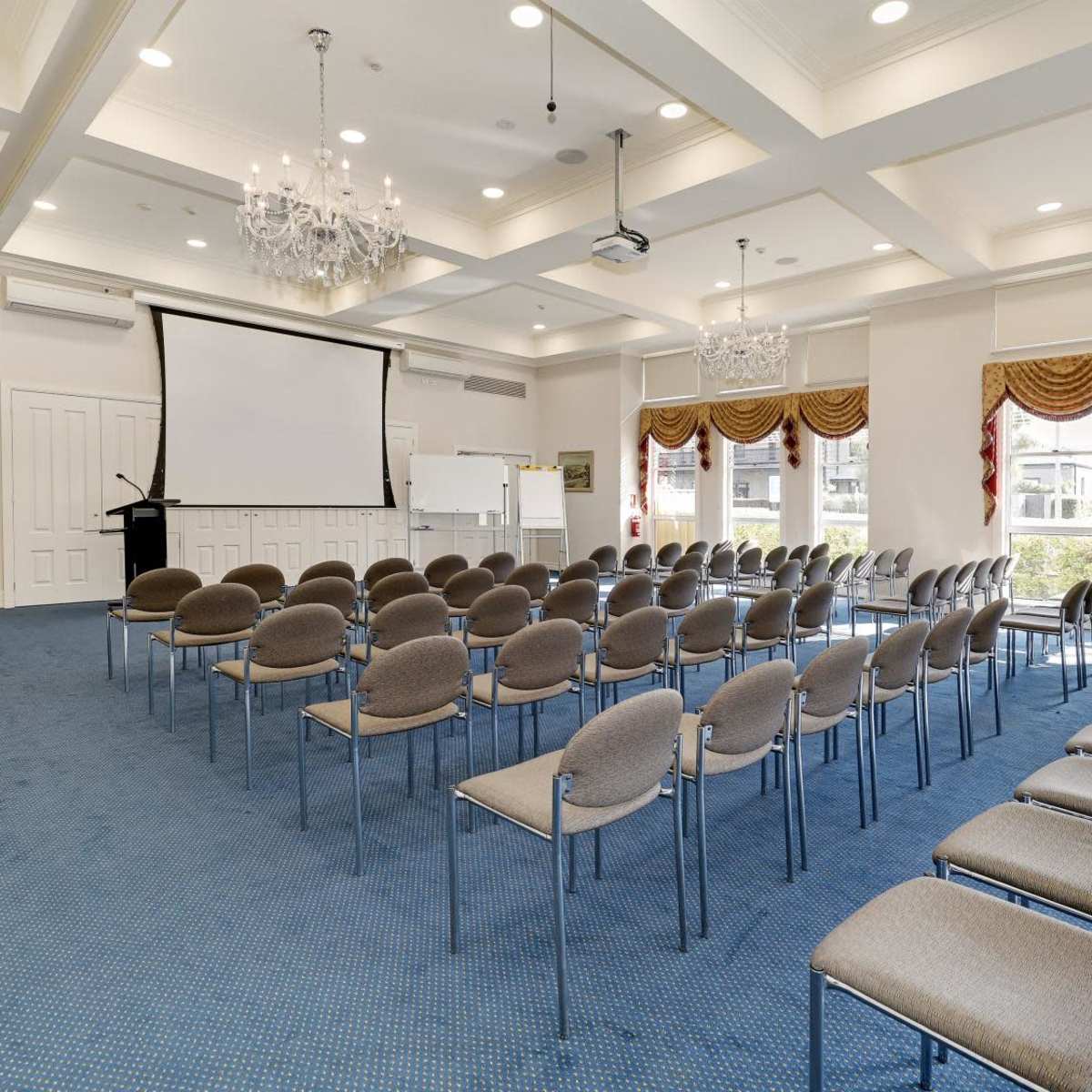 A conference room with rows of chairs facing a projection screen and a podium, featuring a carpeted floor and decorative chandeliers.