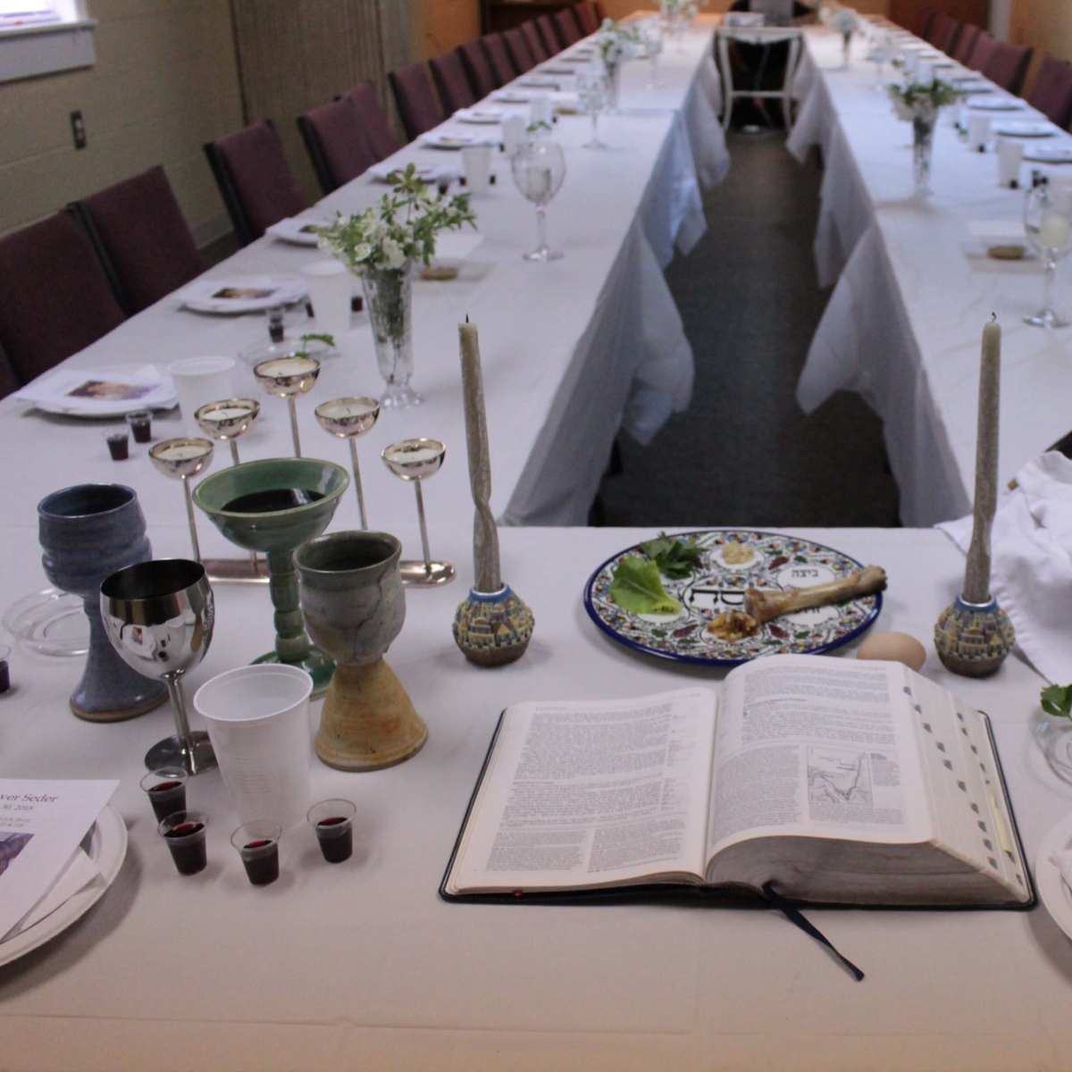 A long dining table is set with white tablecloths, glassware, and a variety of ceremonial items, including a large open book and small cups arranged along the center.