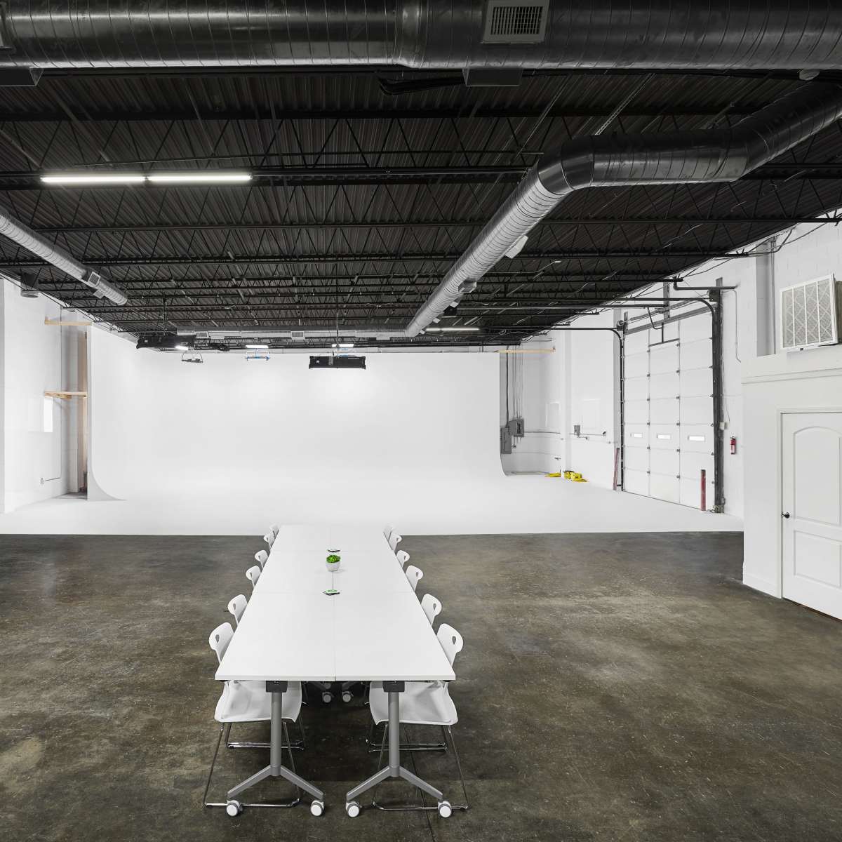 A large, empty studio space with a long white table surrounded by chairs, and a white backdrop at one end.