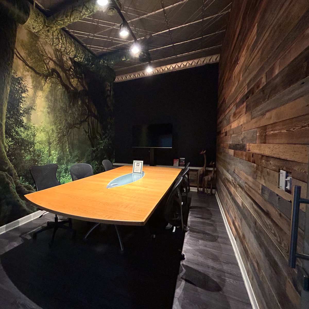 A modern conference room with a large wooden table, black chairs, and a mural of a forest on one wall.