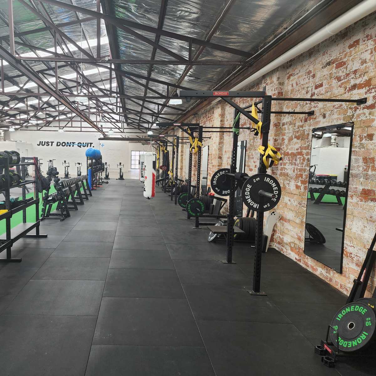 A gym interior with weightlifting equipment, including barbells and kettlebells, arranged along one side and a mirror mounted on the wall, alongside a motivational phrase displayed above.
