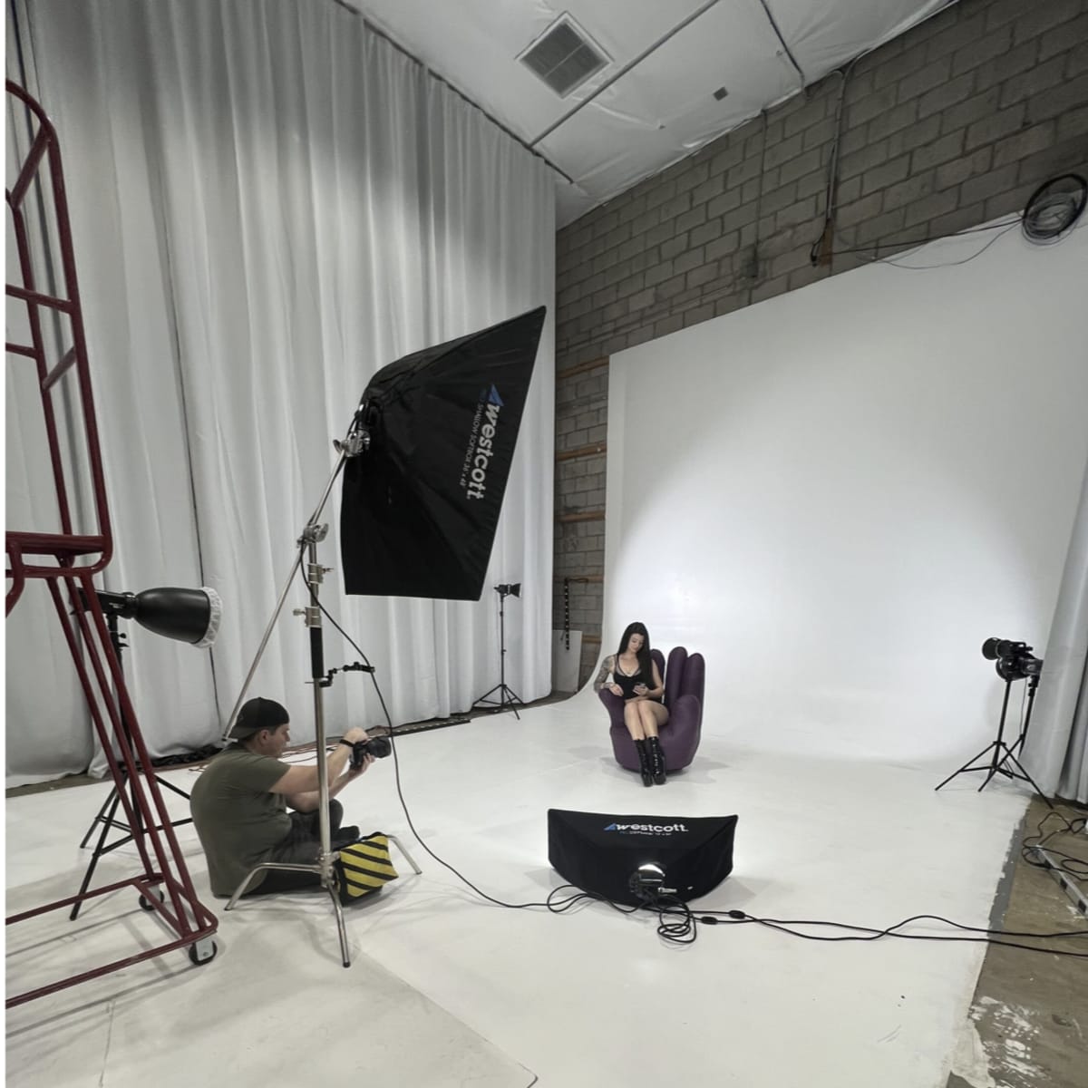 A photoshoot is taking place in a studio, featuring a model sitting on a chair while a photographer captures the scene with studio lighting equipment set up around them.
