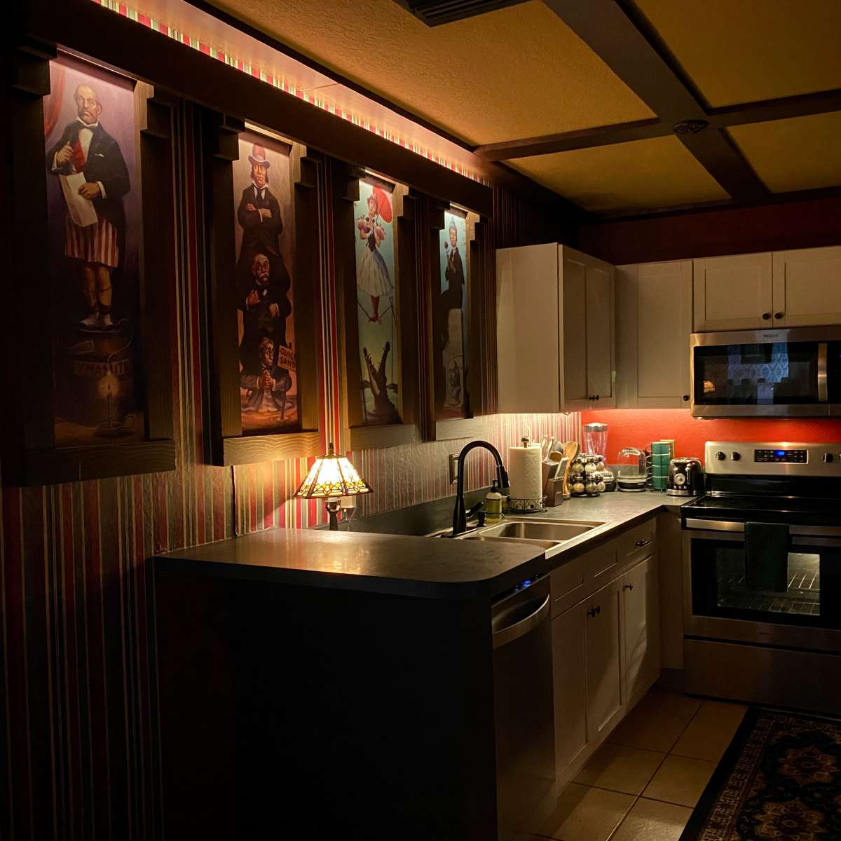 A dimly lit kitchen featuring white cabinets, stainless steel appliances, and artwork on the walls.