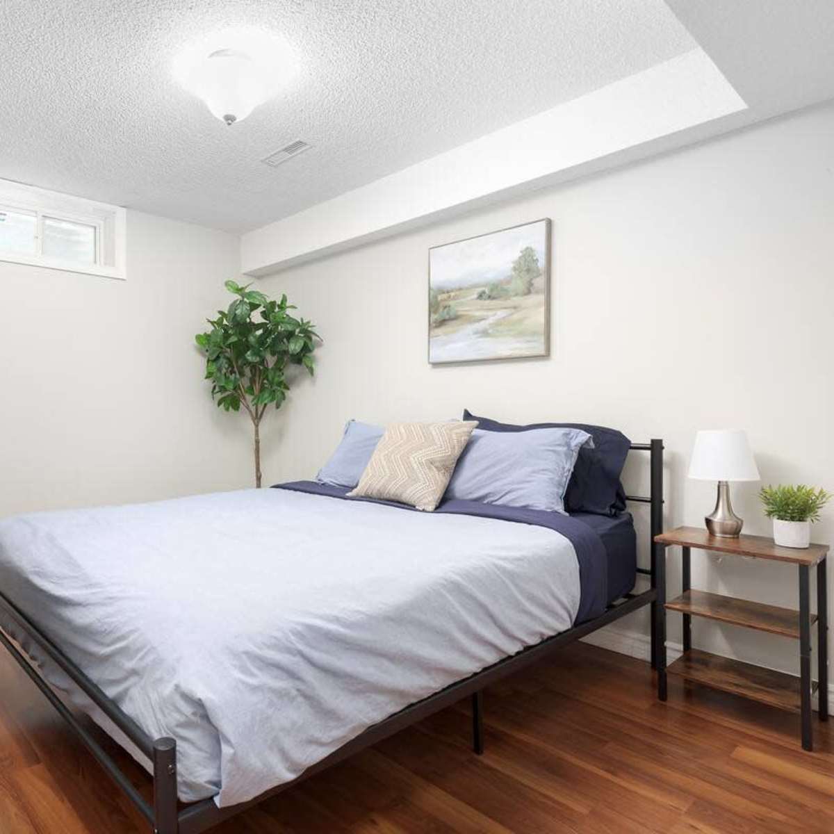 A small bedroom with a bed, a nightstand, a plant, and a dresser against a light-colored wall.