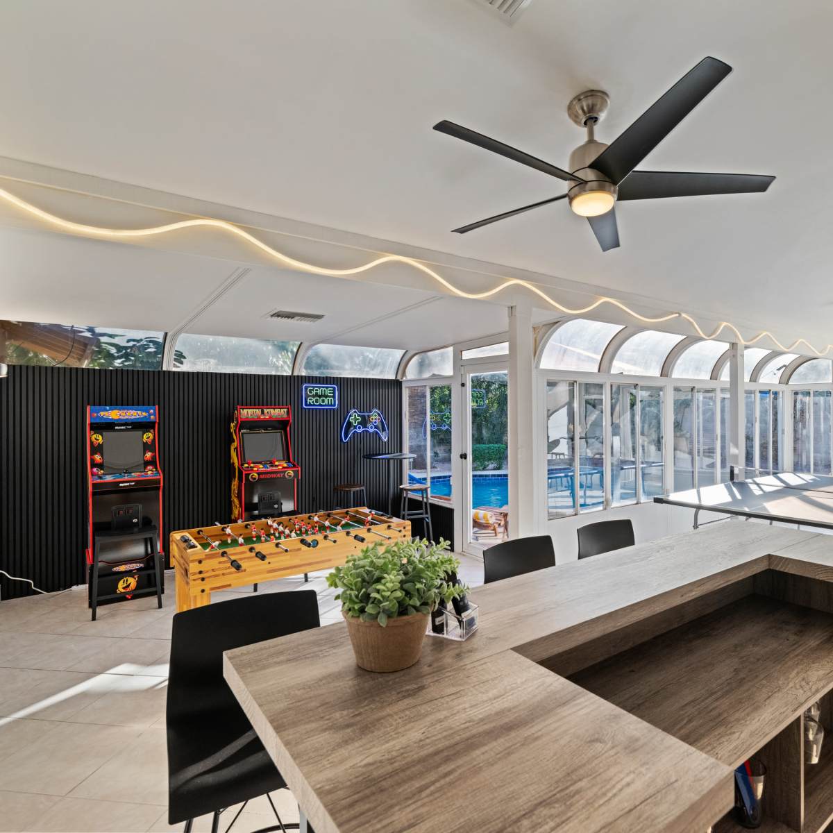 A modern game room featuring arcade machines, a foosball table, and a view of a pool through large windows.