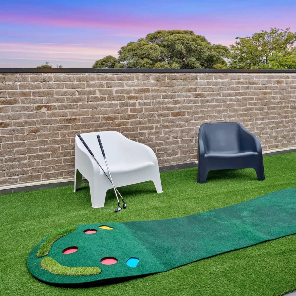 A putting green with colorful holes, two modern chairs, and golf clubs resting against a brick wall under a colorful sky.