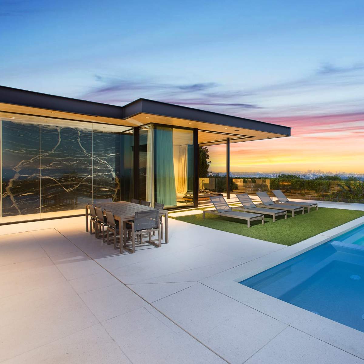 A modern house features a sleek pool and dining area with lounge chairs, overlooking a vibrant sunset.