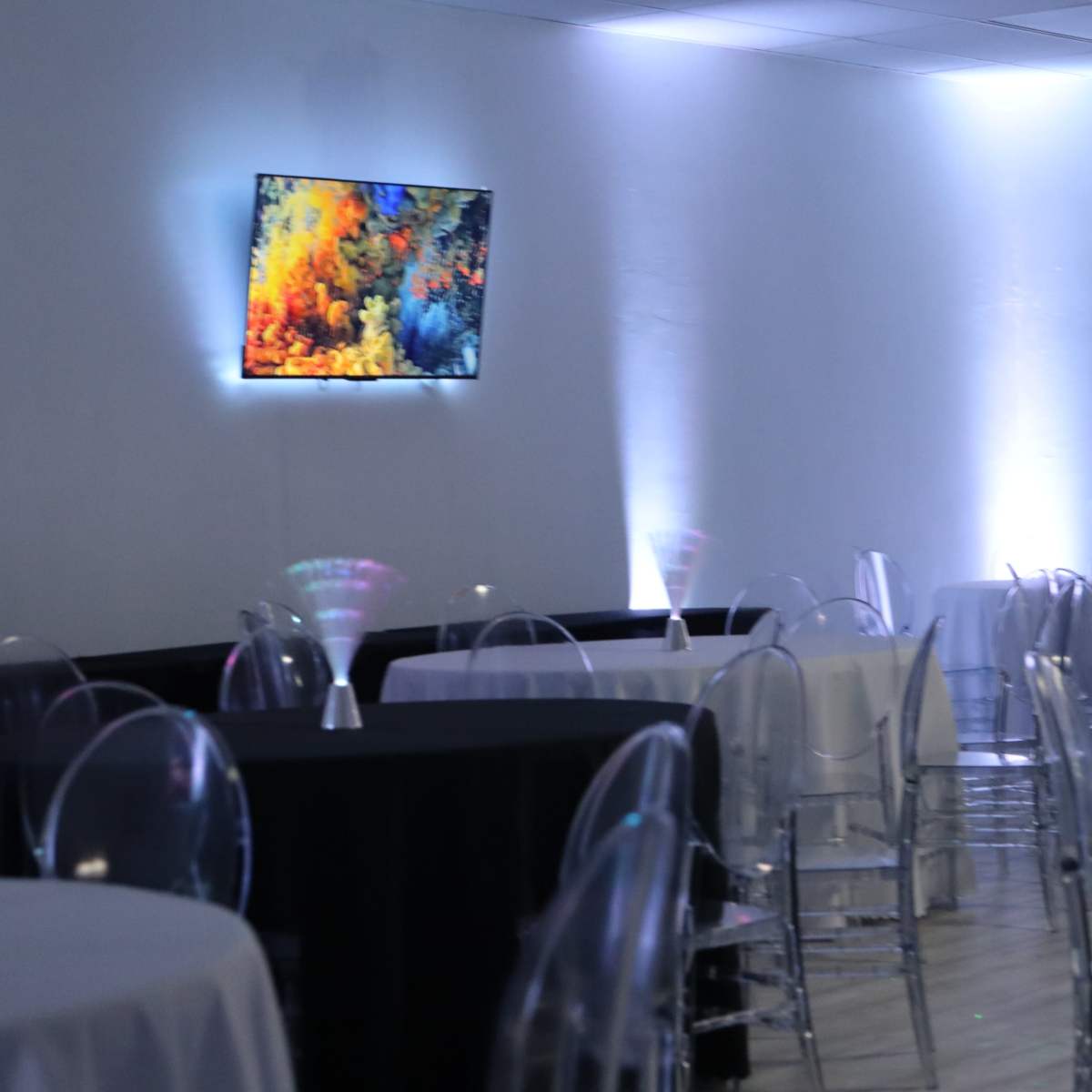A spacious event room features several round tables with black and white tablecloths, transparent chairs, and a colorful artwork displayed on the wall.