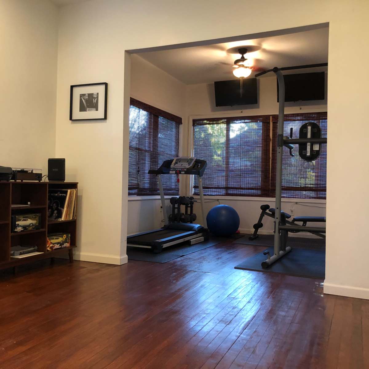 A home gym area with exercise equipment, including a treadmill and a weight bench, adjacent to a living space featuring a bookshelf and a printer.
