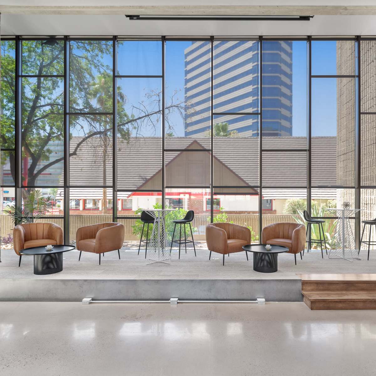 A modern seating area with brown chairs and small tables, facing large windows that overlook a cityscape and a shaded outdoor space.