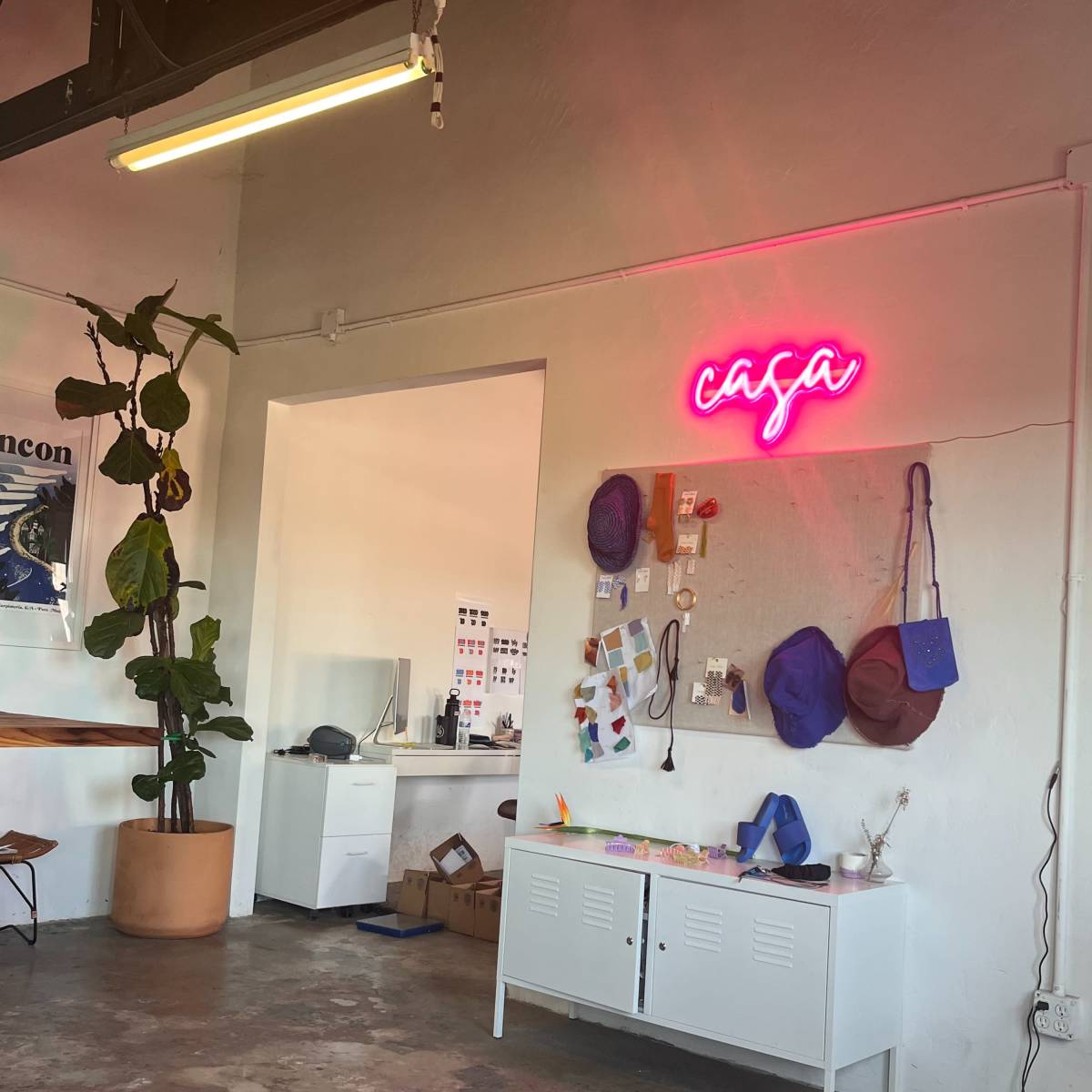 A minimalist interior featuring a white cabinet, a neon sign that reads "casa," a potted plant, and various accessories hung on the wall.
