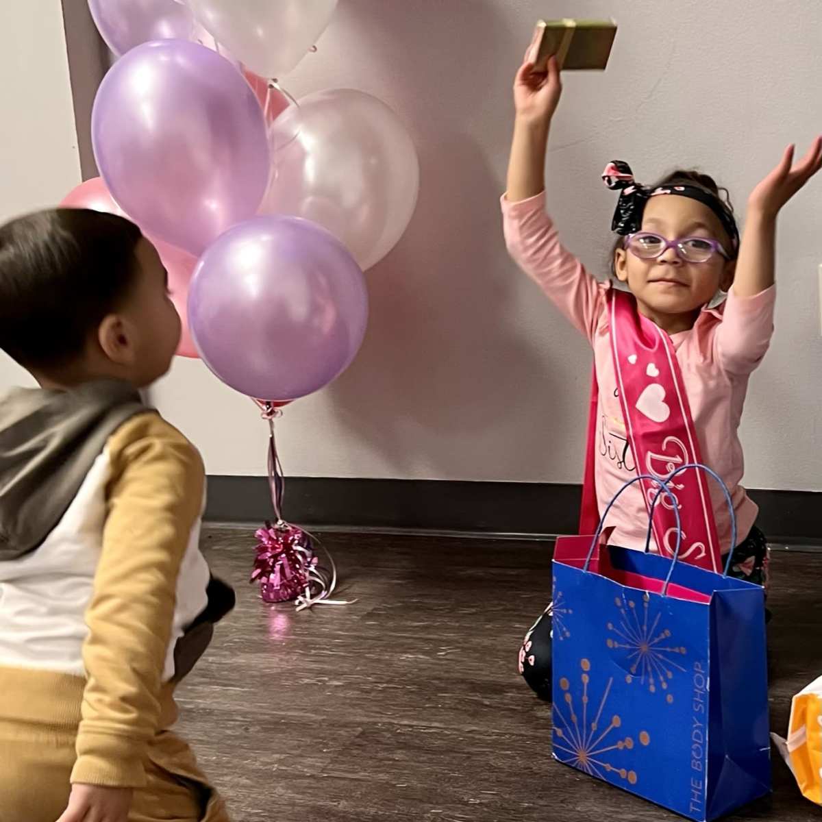 A girl wearing a pink shirt and glasses joyfully holds up a small gift while a boy in a brown and white outfit watches near a cluster of balloons.
