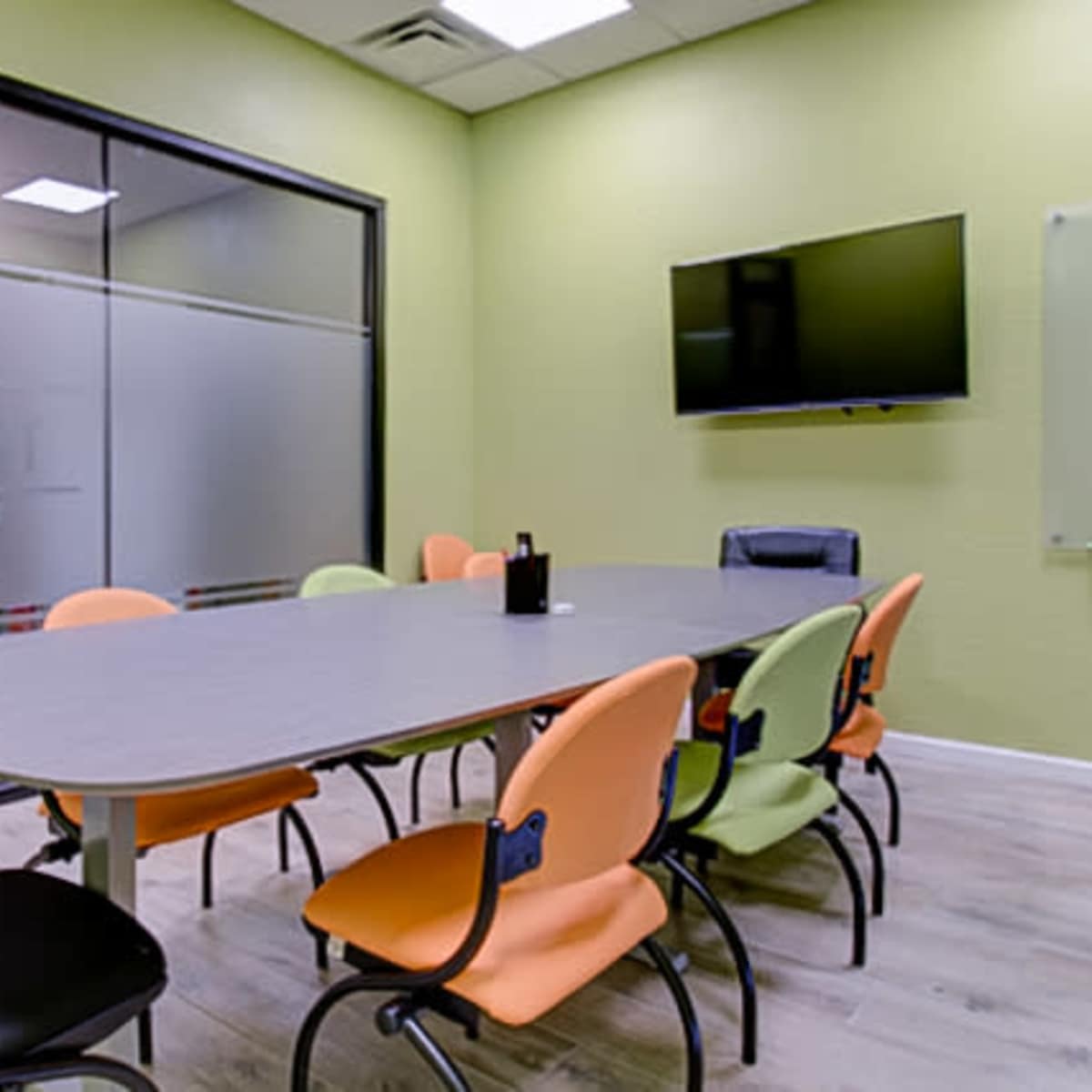 A meeting room features a large table surrounded by six chairs in various colors, with a television mounted on the wall and a glass partition showing the room's name.