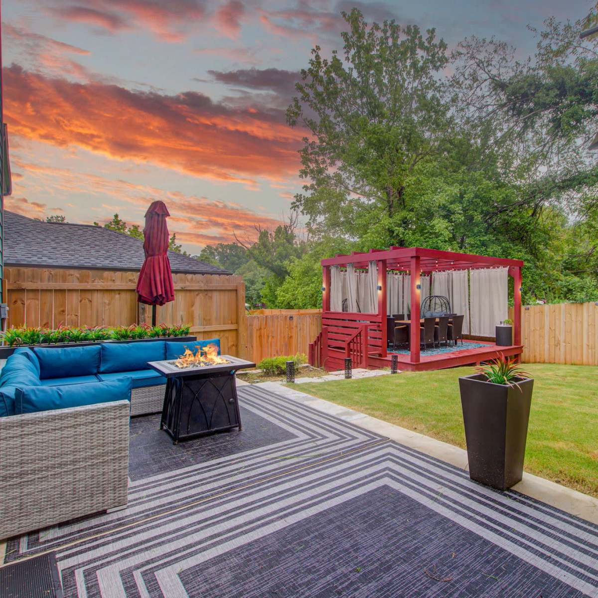 A backyard patio with a blue couch, a black coffee table, and a red gazebo against a backdrop of a colorful sunset.