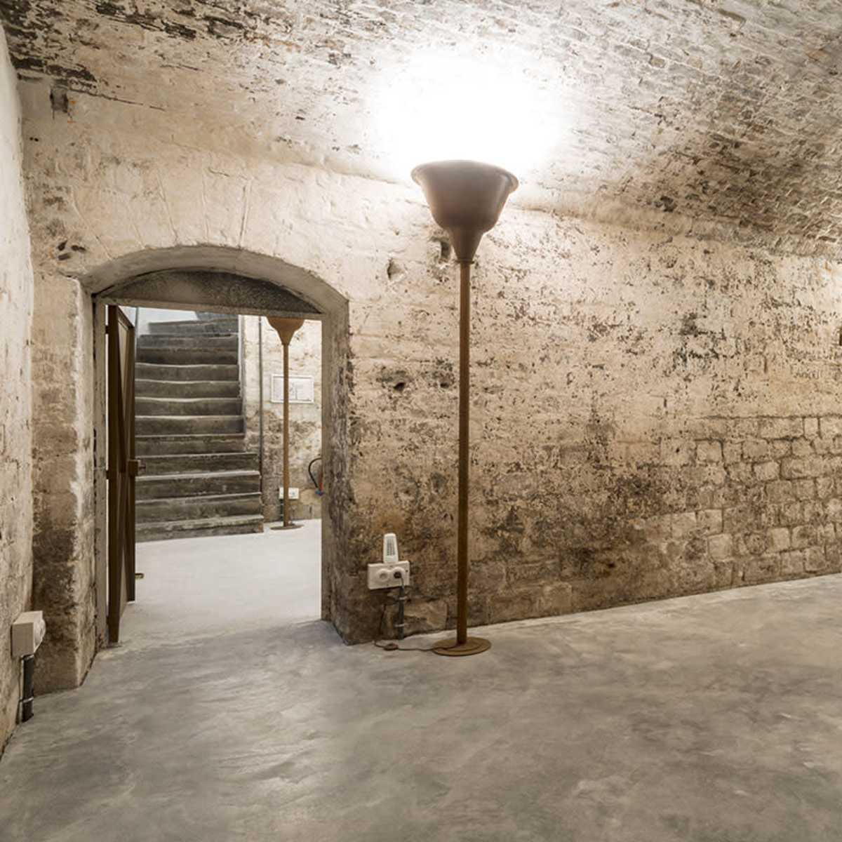 An empty basement space with rough stone walls, two standing lamps, and a staircase leading to an upper level.