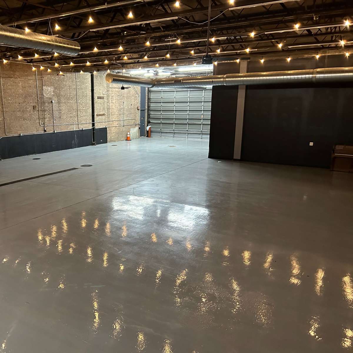 A spacious, industrial-style room with a polished concrete floor and exposed brick walls, featuring overhead lighting and a large garage door at one end.