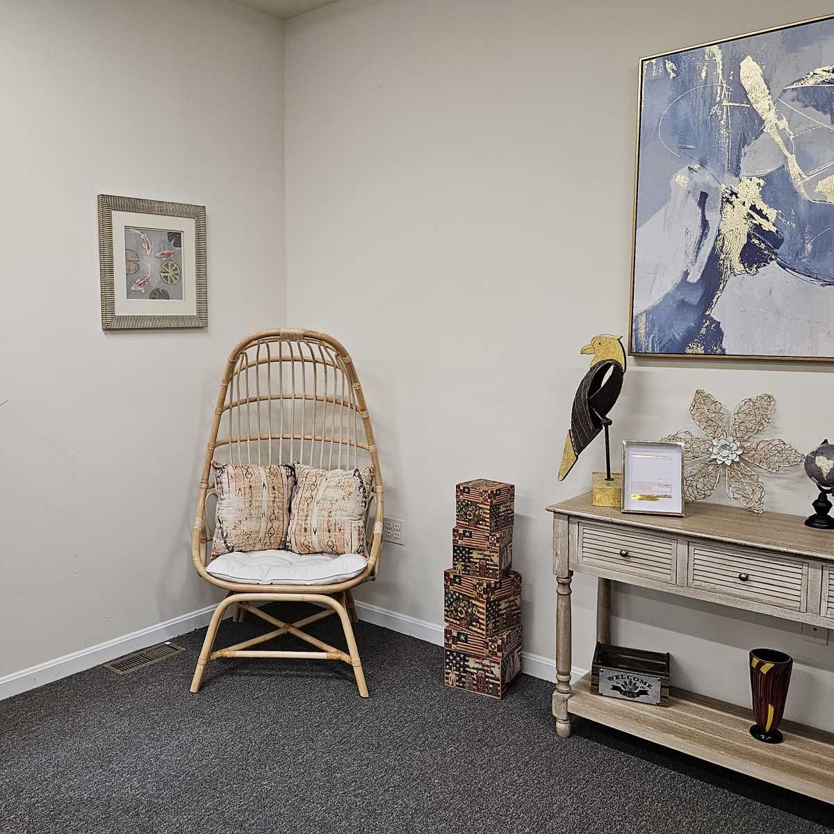 A cozy corner features a rattan chair with cushions beside a window, accompanied by decorative plants and a small table adorned with various items.