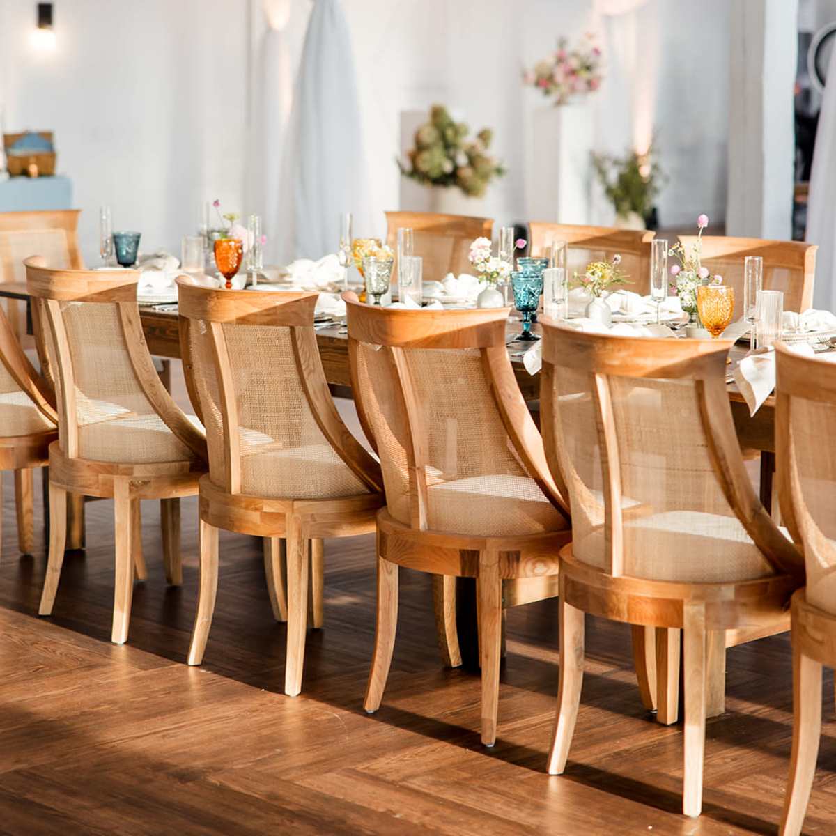 A long wooden dining table is set with white tableware and surrounded by multiple wooden chairs in a decorated event space with soft lighting and draped fabric.