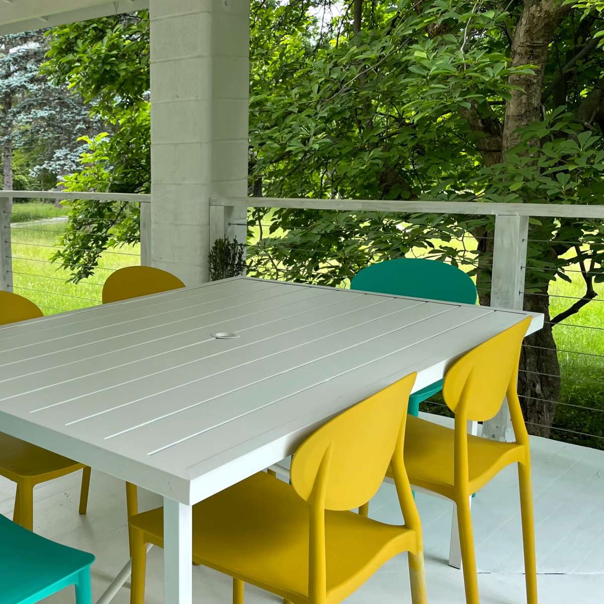 A white table with six brightly colored chairs is arranged on a covered porch overlooking a green lawn and trees.