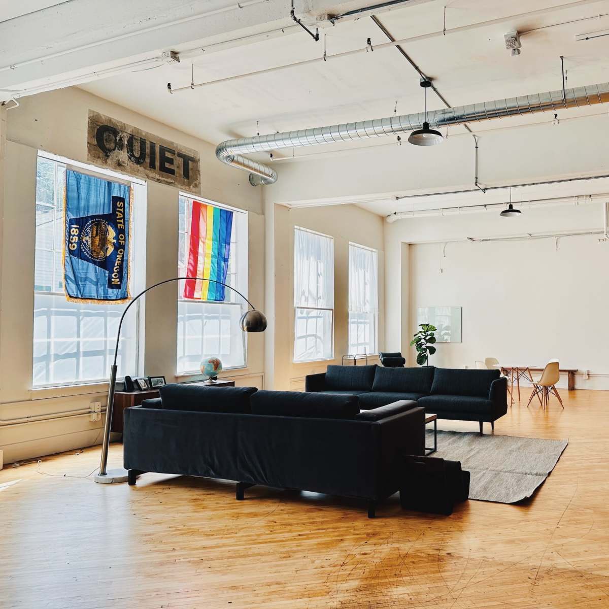 A spacious, minimalist room with wooden floors, featuring two black sofas, a large floor lamp, a colorful flag and a sign that says "QUIET," along with a dining area in the background.