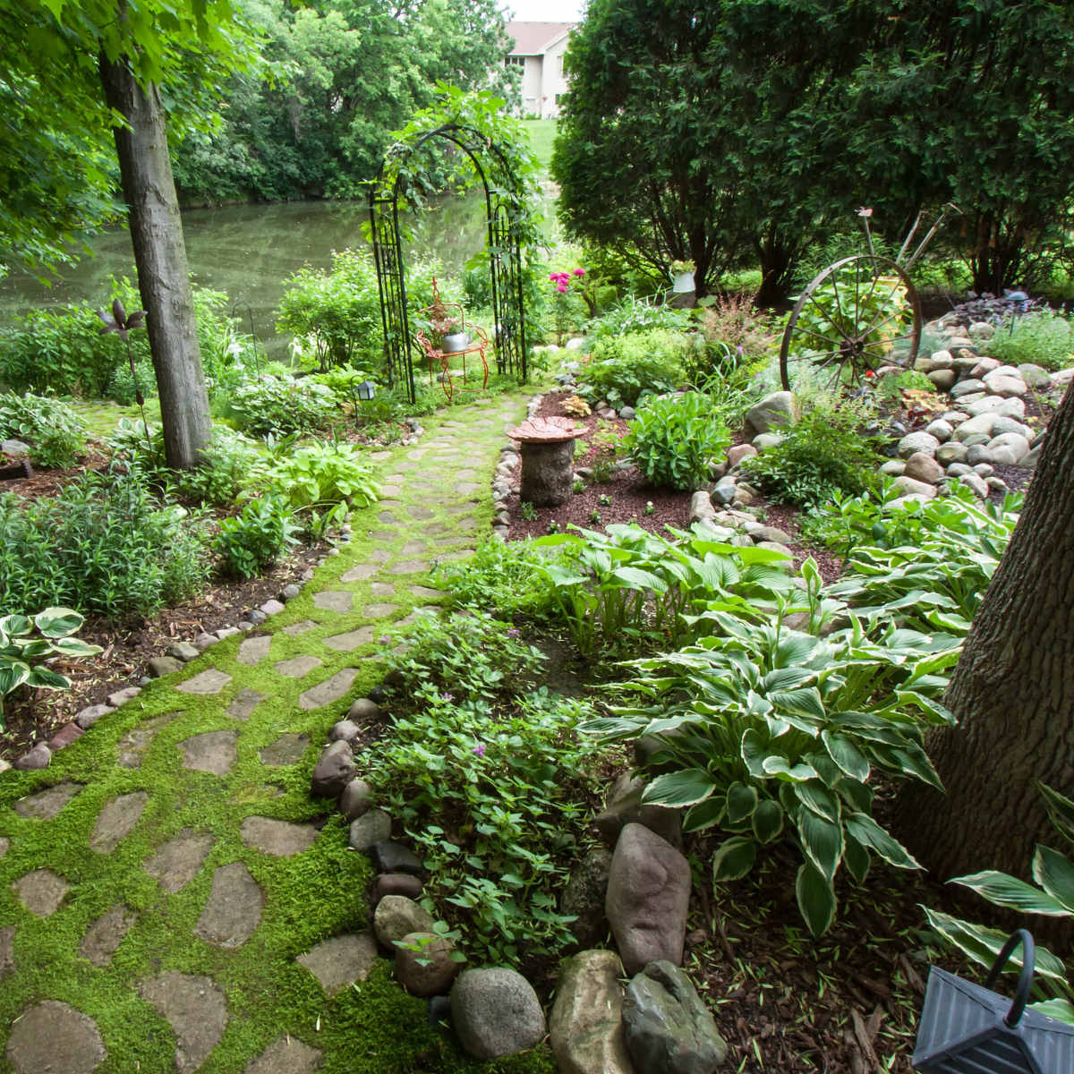 A stone pathway winds through a lush garden, leading to an archway and a quiet waterway in the background.