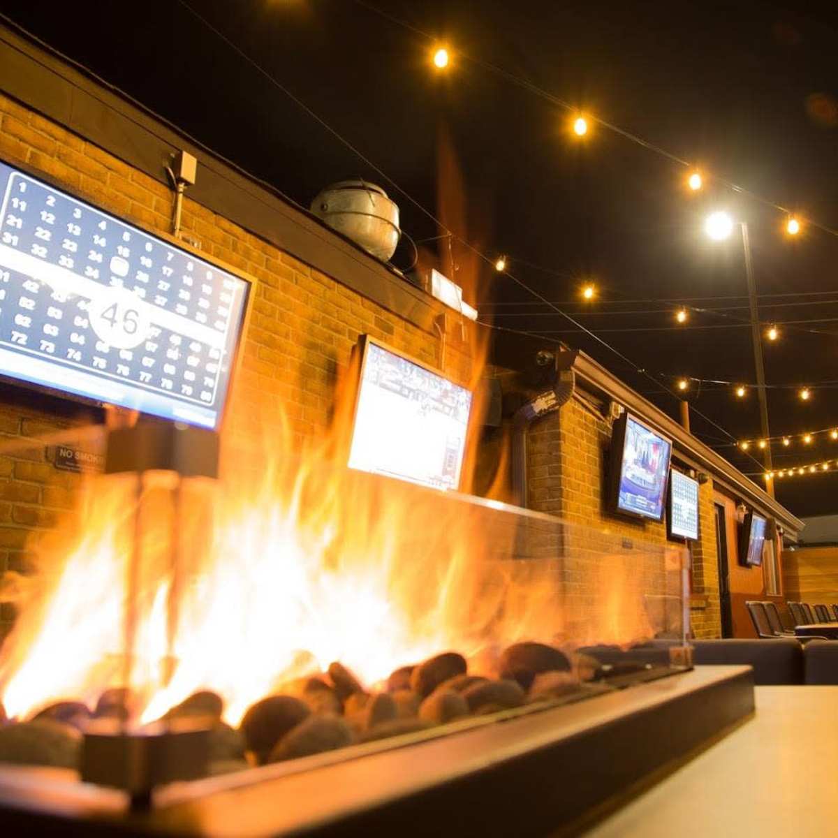 An outdoor seating area with a central fire feature, several TVs mounted on the walls, and string lights overhead.