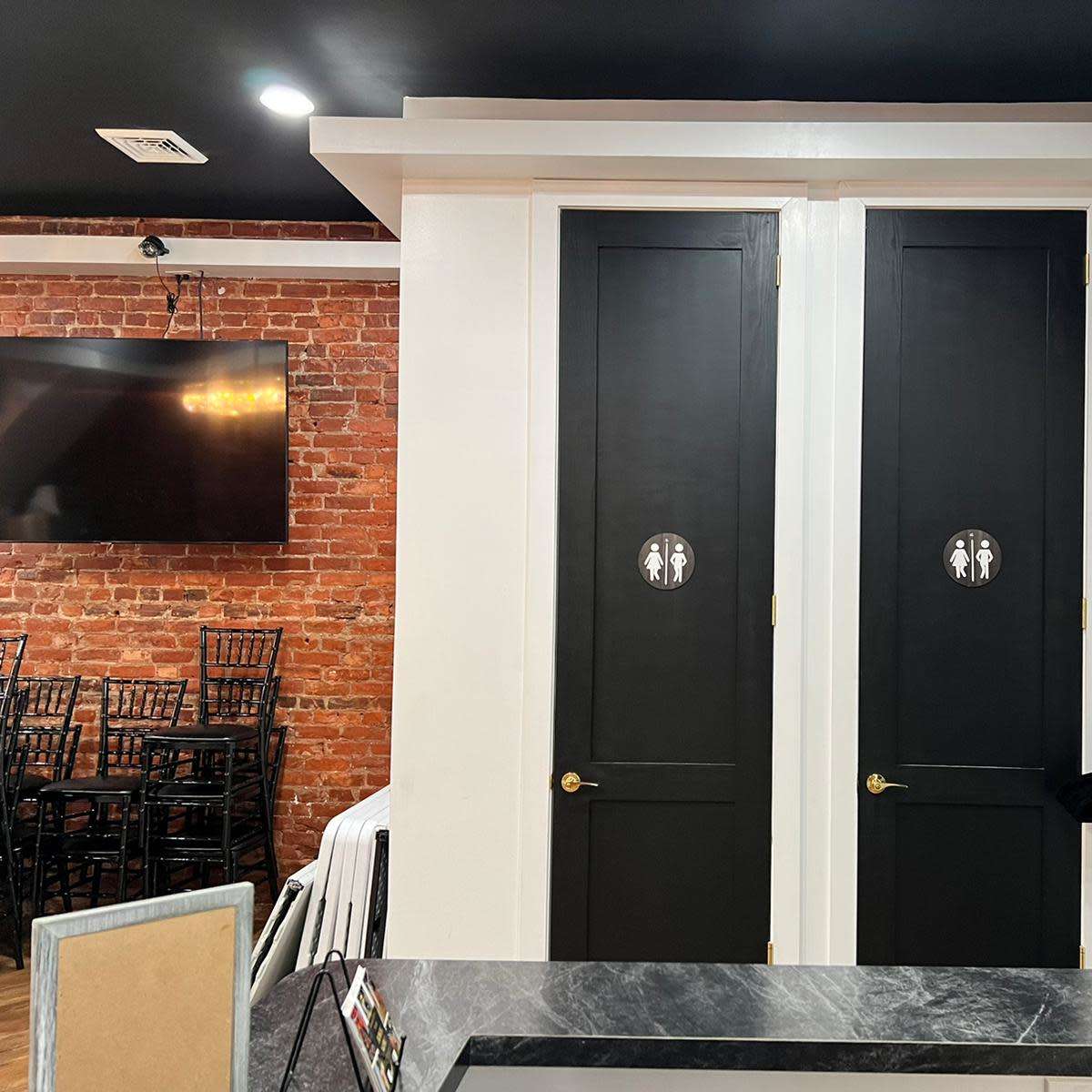 A restroom area featuring two black doors, each with a circular bathroom sign, next to a dark brick wall and a television mounted on the opposite wall.