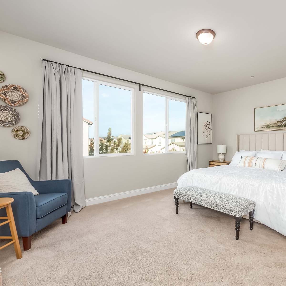 The bedroom features a large bed with white linens, a bench at the foot, a seating area with a blue chair, and windows providing natural light.