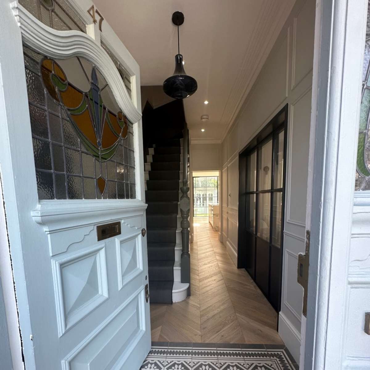 An open front door revealing a bright hallway with a staircase, patterned tile flooring, and a view of a glass-paneled door at the end.