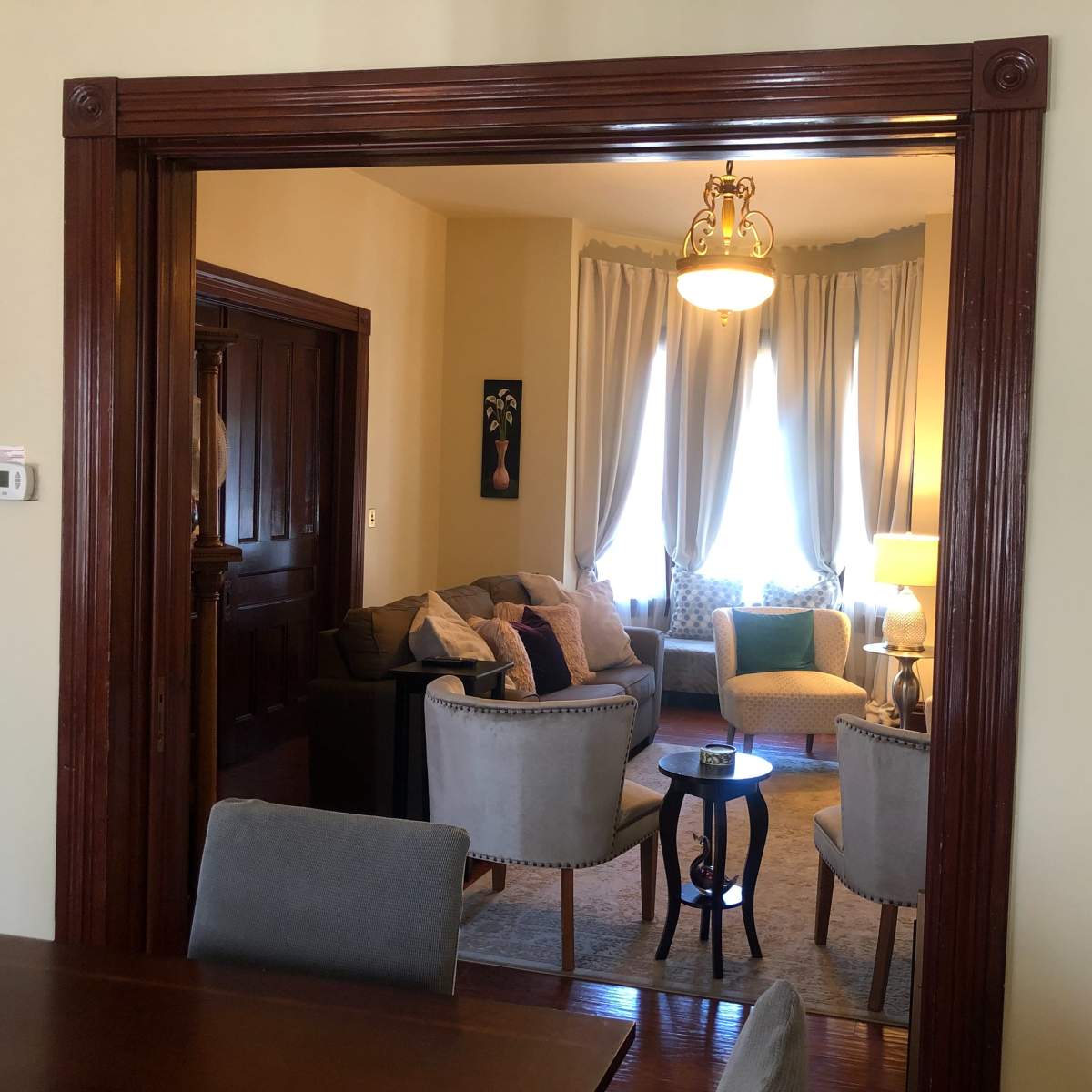 A view through an architectural archway into a living room furnished with a sofa, side chairs, and a lamp, illuminated by natural light from large windows.