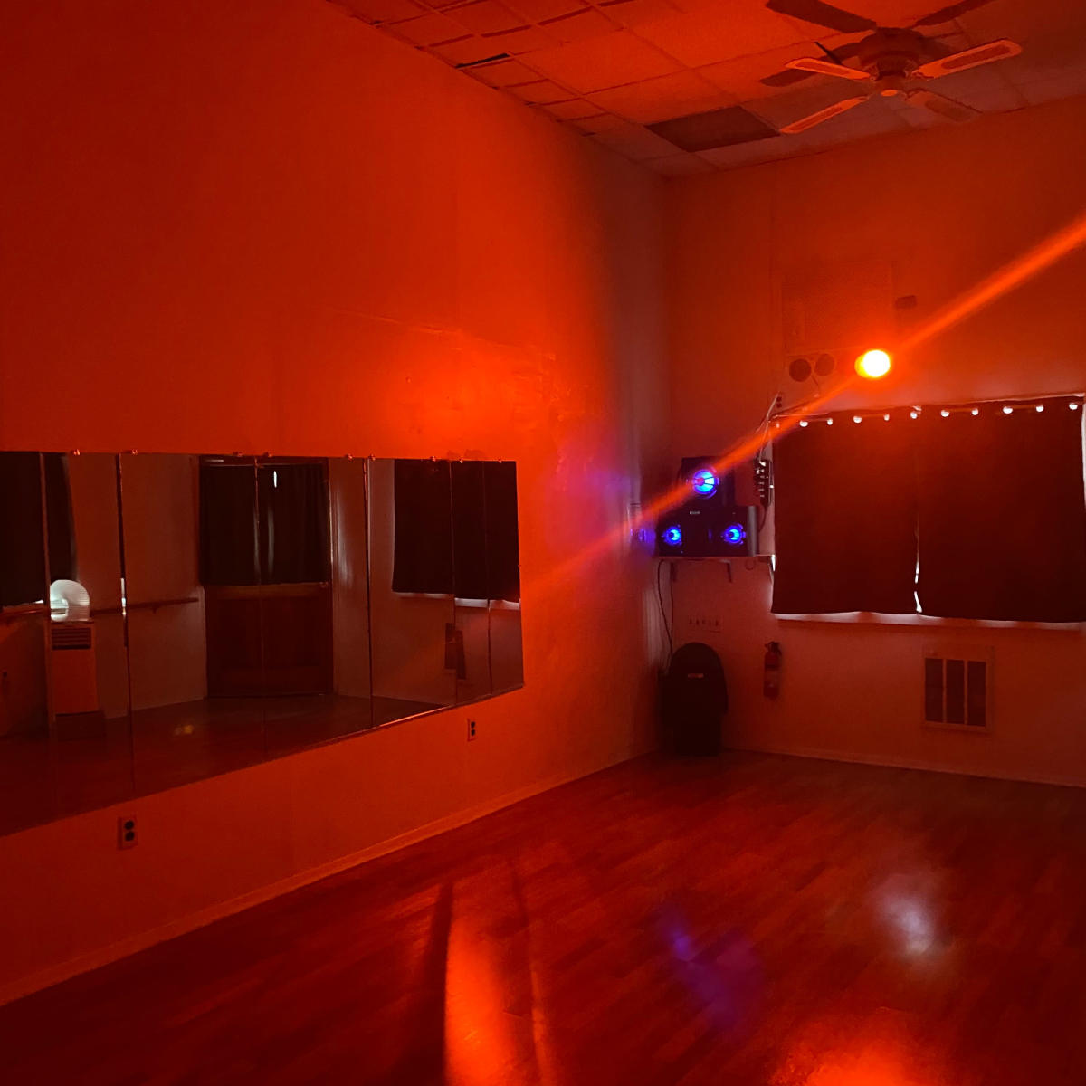 The interior of a dimly lit dance studio features a mirror along one wall, a window with dark curtains, and a corner illuminated by a single orange light.