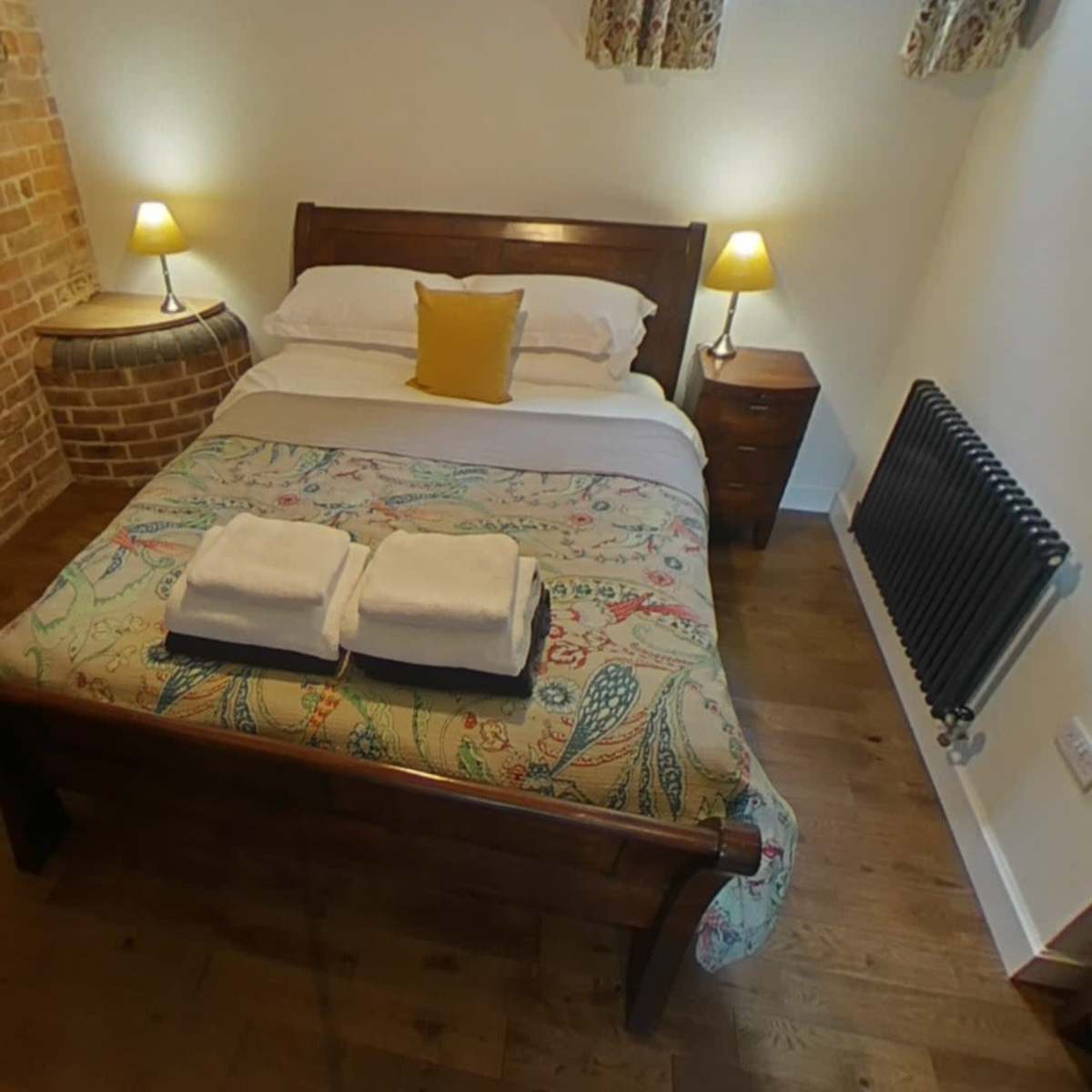A small bedroom featuring a wooden bed with a patterned bedspread, two nightstands with lamps, and a brick wall.