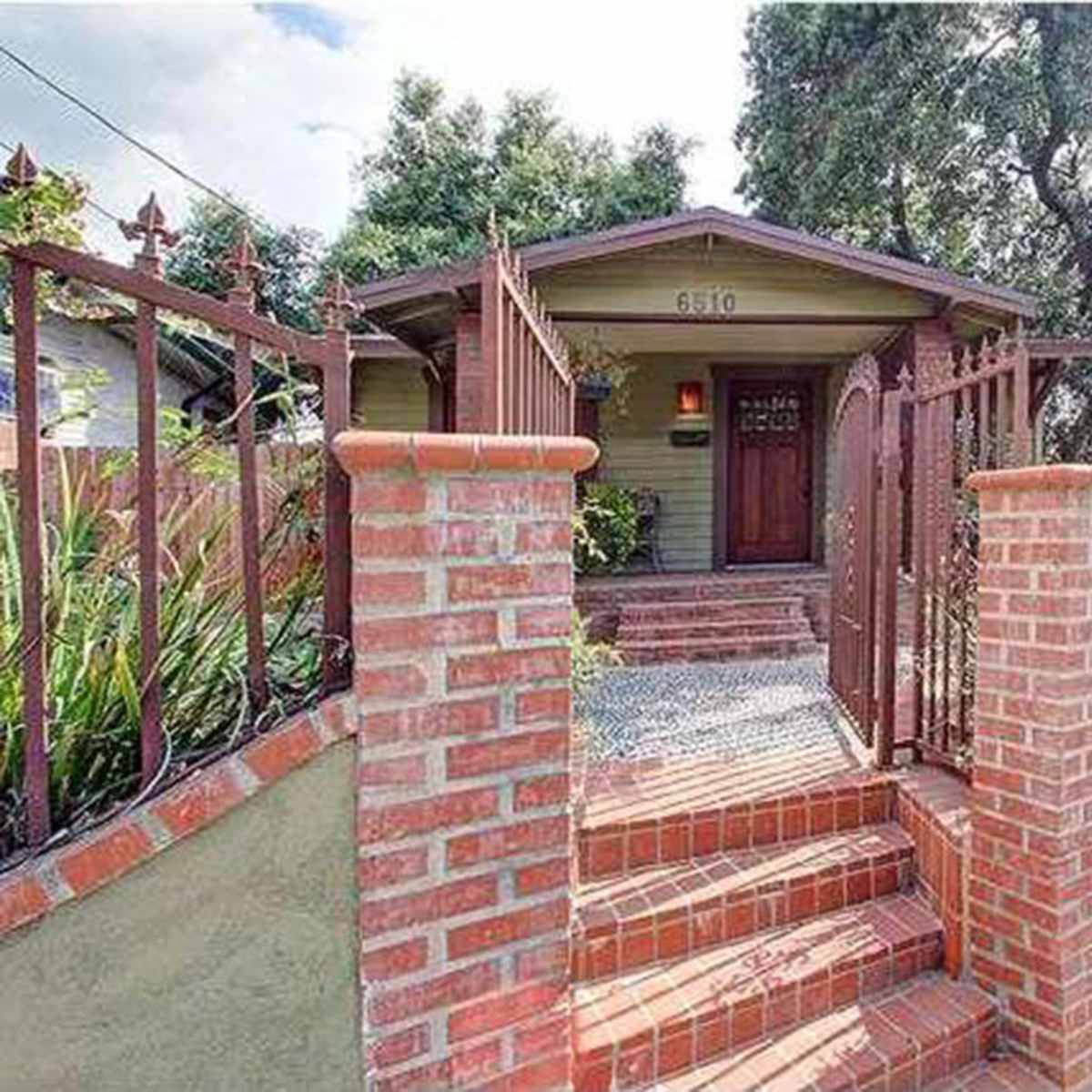 A small house with a brick walkway and decorative fencing is set behind a low wall and surrounded by greenery.