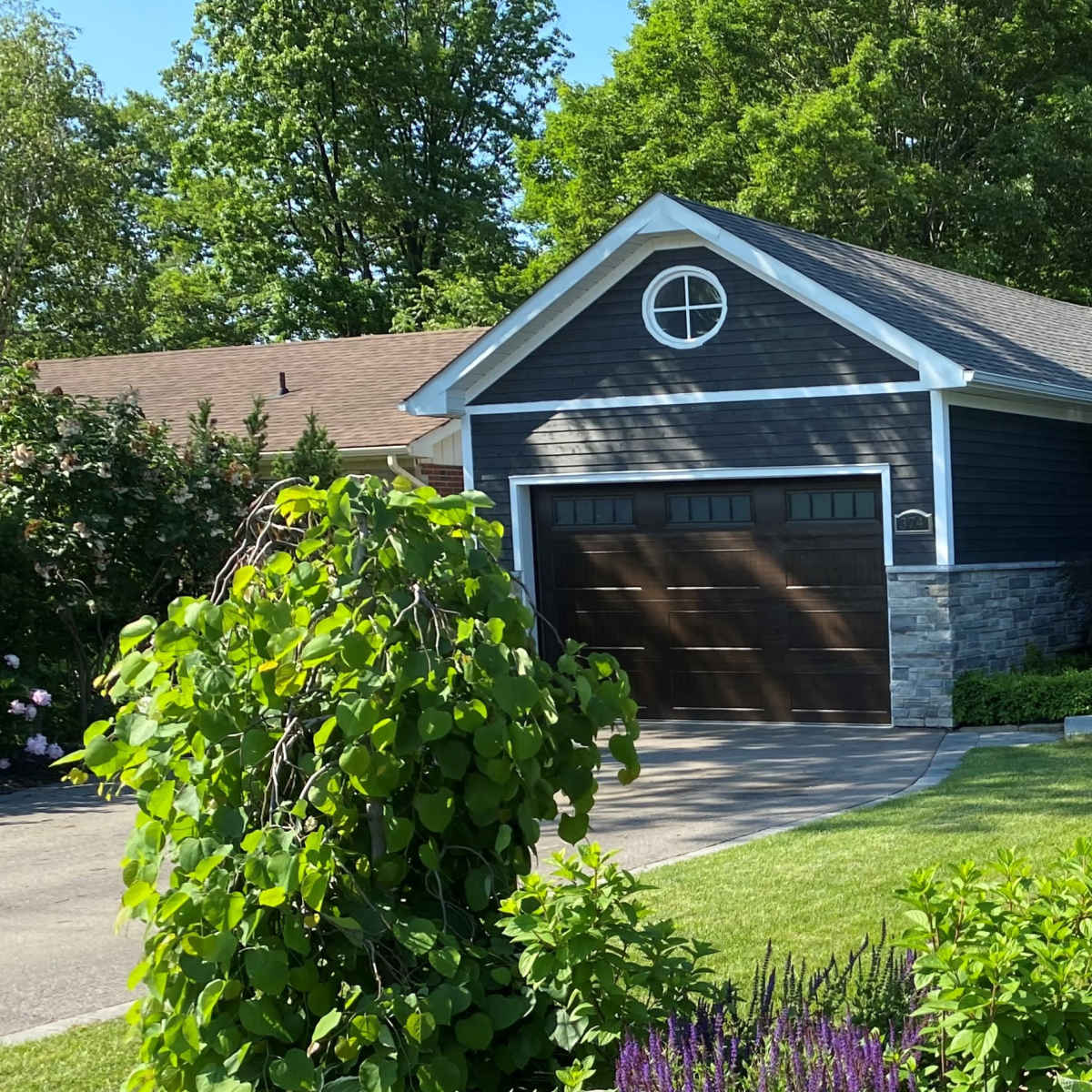 A single-story house with a dark exterior, a stone veneer, and a two-car garage is surrounded by green lawns and landscaping.