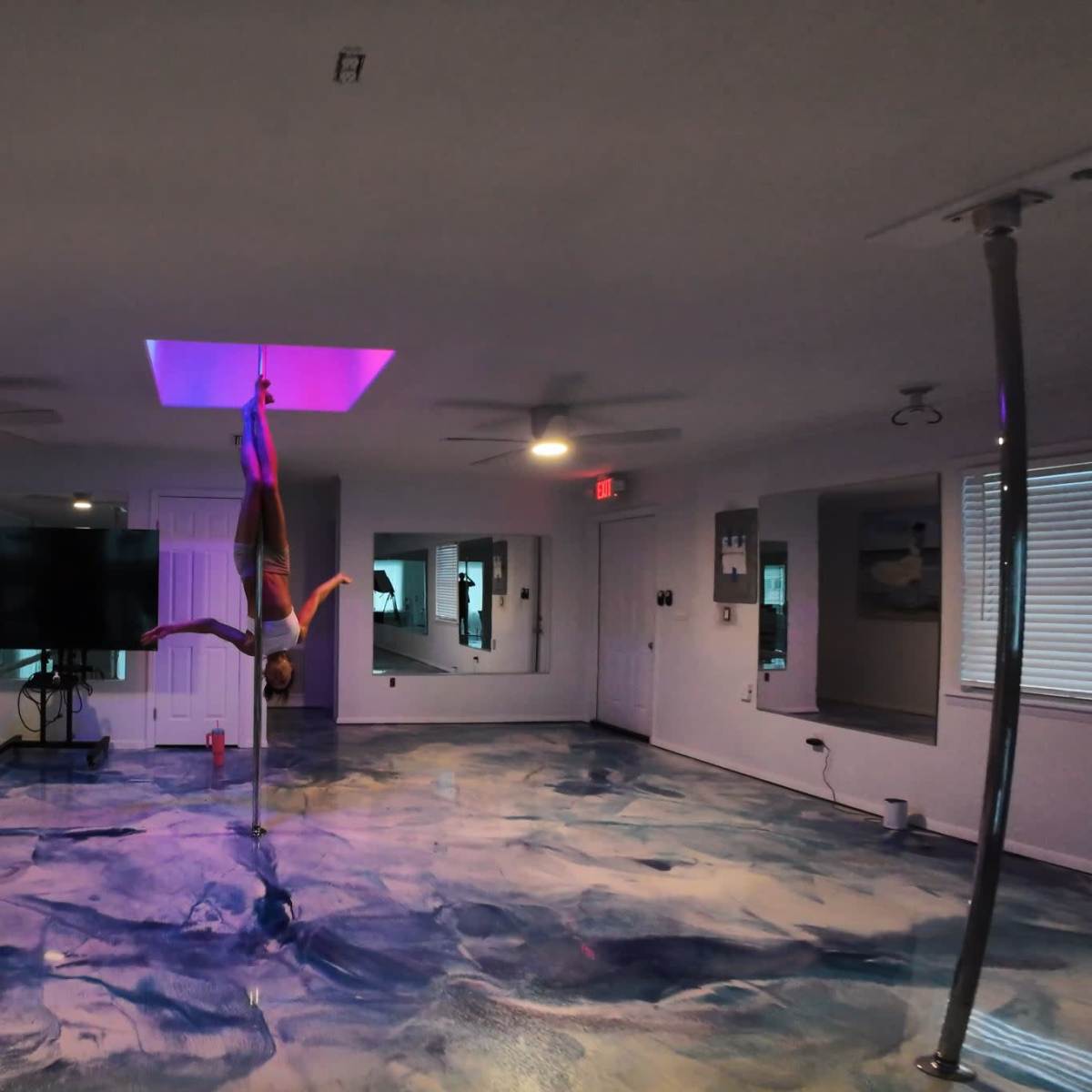 A person performs on a pole in a brightly lit room with an artistic floor design and multiple windows.