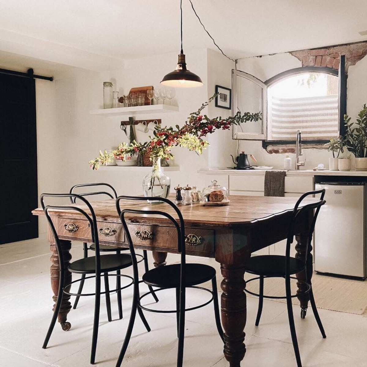 A rustic kitchen features a wooden table surrounded by black chairs, with a vase of flowers and various kitchen appliances visible in the background.