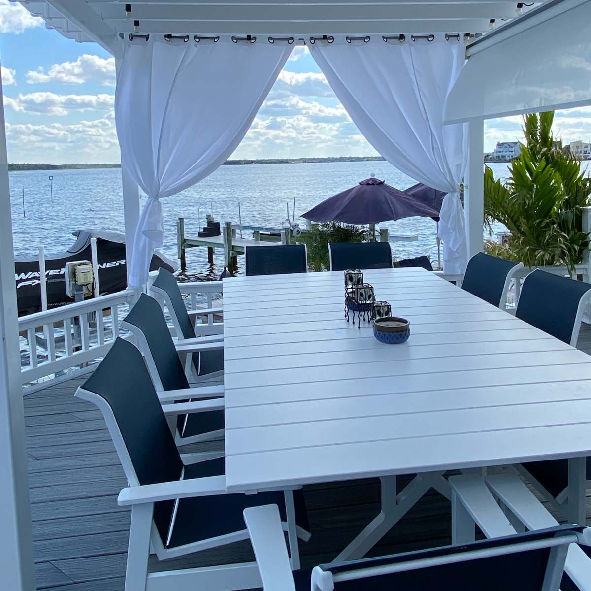 A white table with black chairs is set on a deck overlooking a body of water, framed by flowing white curtains.