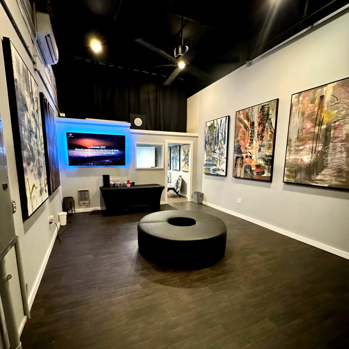 A modern gallery space featuring a round seating area in the center, with large abstract paintings on the walls and a television screen displaying visuals at the far end.