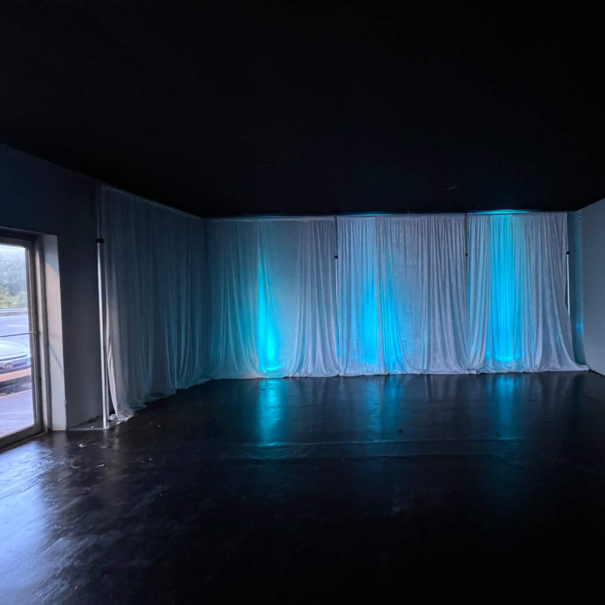 The room is dimly lit with textured black flooring and sheer white curtains backlit in blue along the walls.