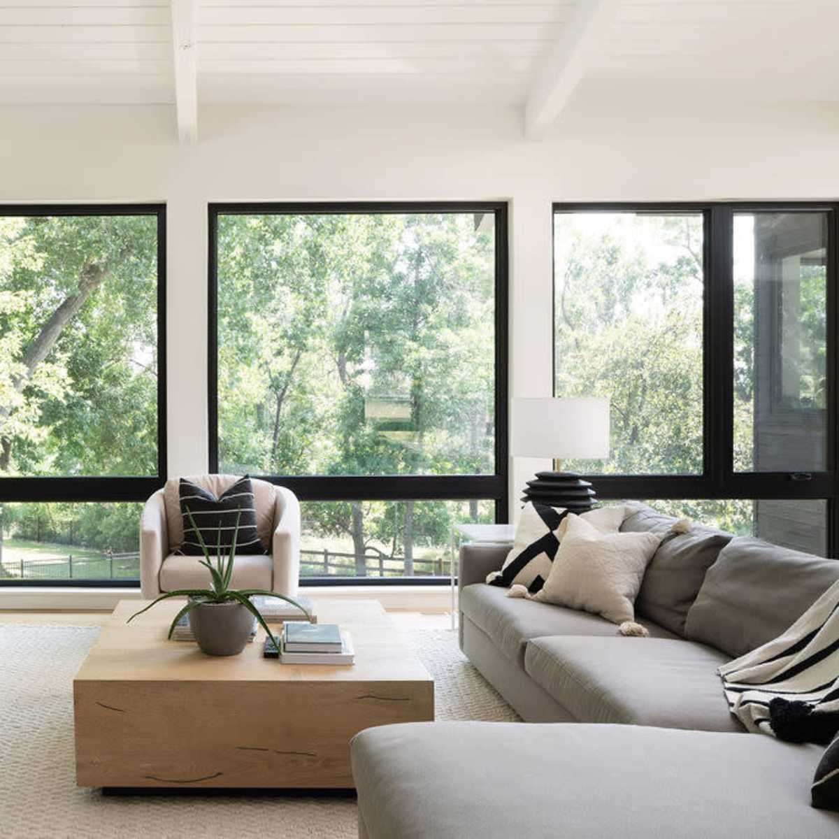 A modern living room featuring large windows that provide a view of greenery, a light-colored sofa, a coffee table, and a potted plant.