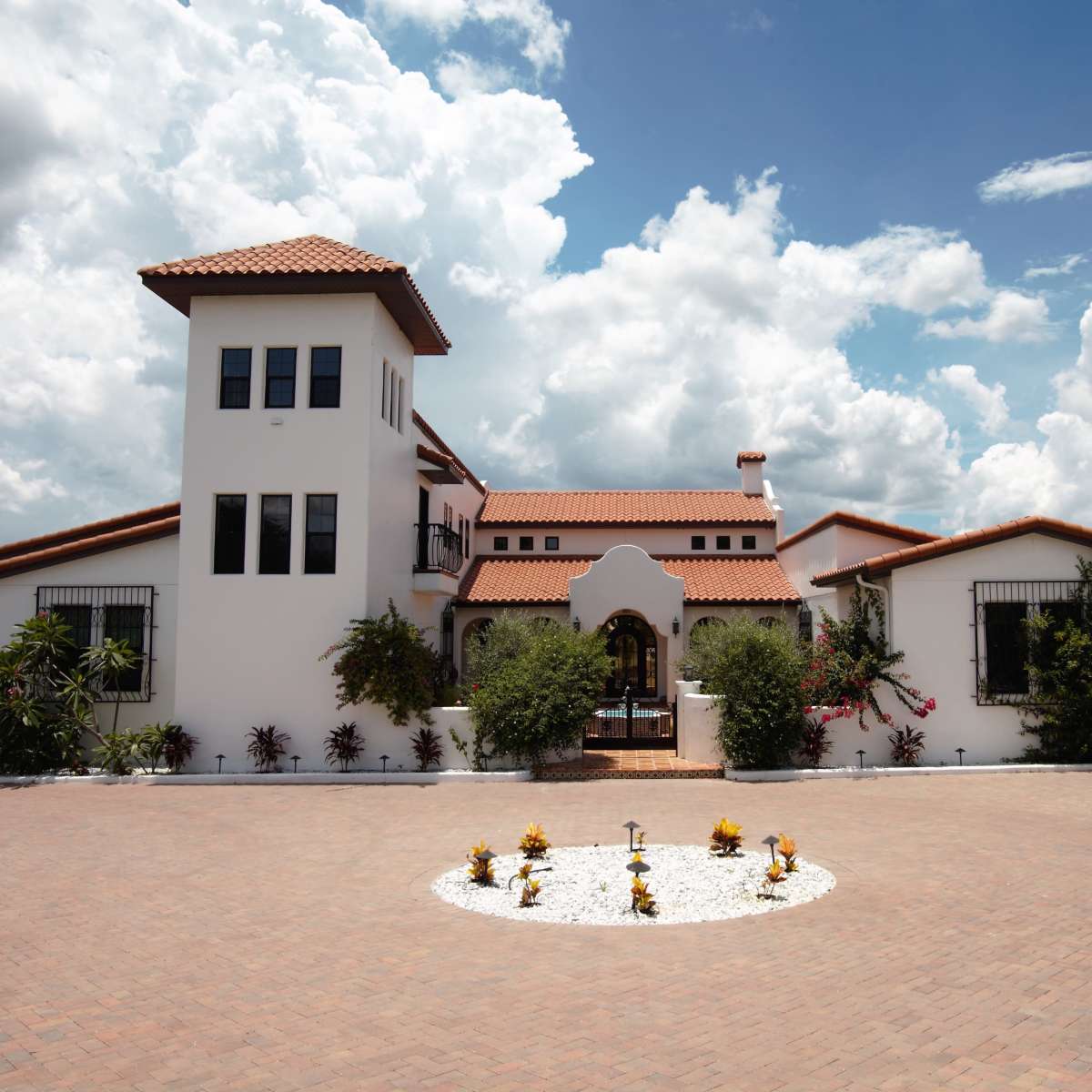 A large, white, two-story house with a red-tiled roof and a tower stands in a landscaped area under a cloudy sky.