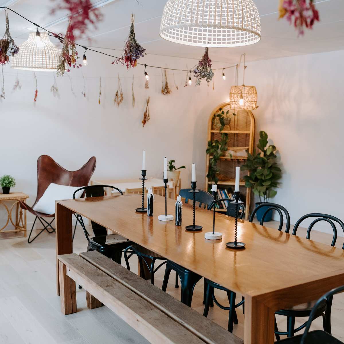 A spacious dining area features a large wooden table surrounded by black chairs and decorated with candles, while hanging plants and dried flowers adorn the walls and ceiling.