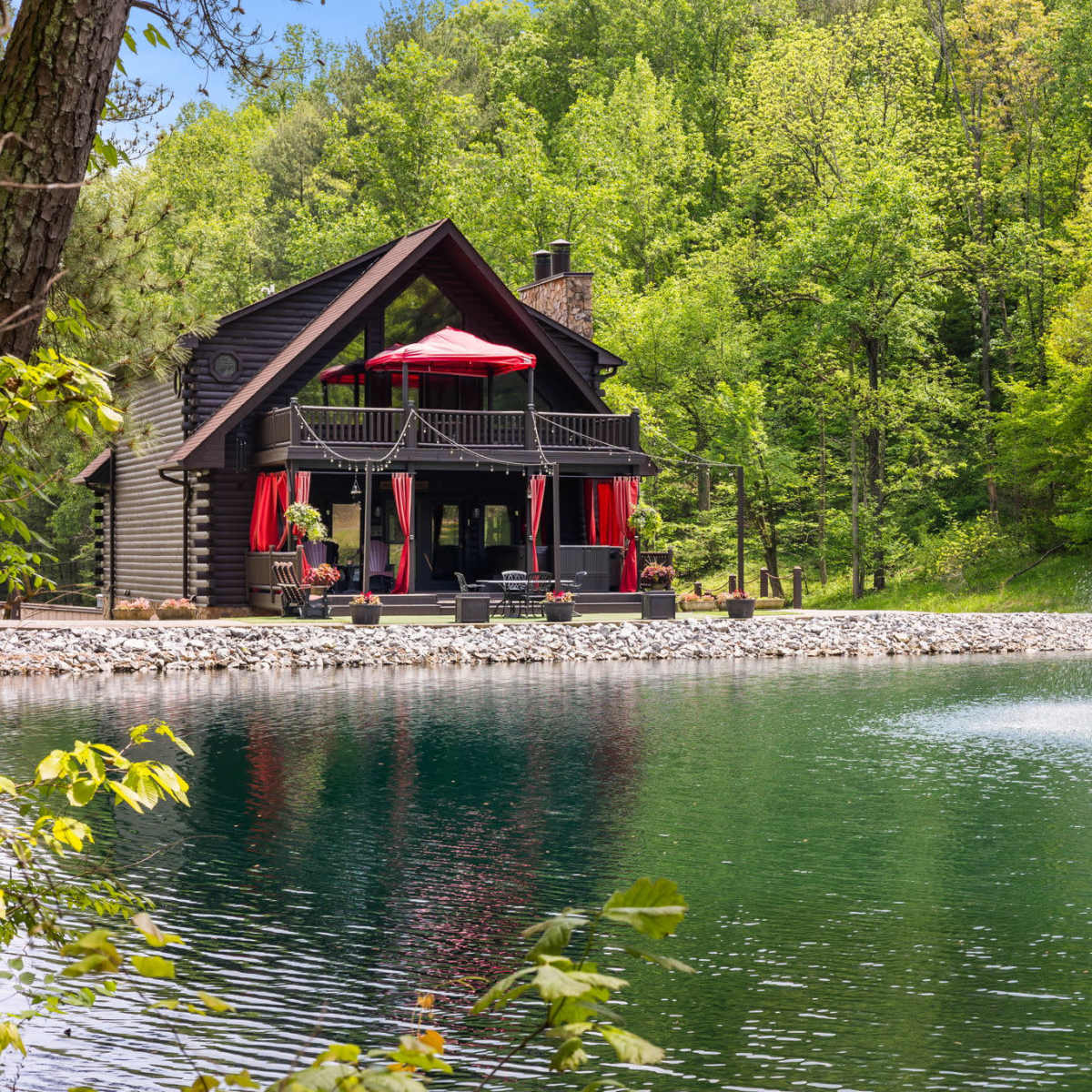 A wooden cabin with red awnings and a deck overlooks a pond featuring a fountain, surrounded by lush green trees.