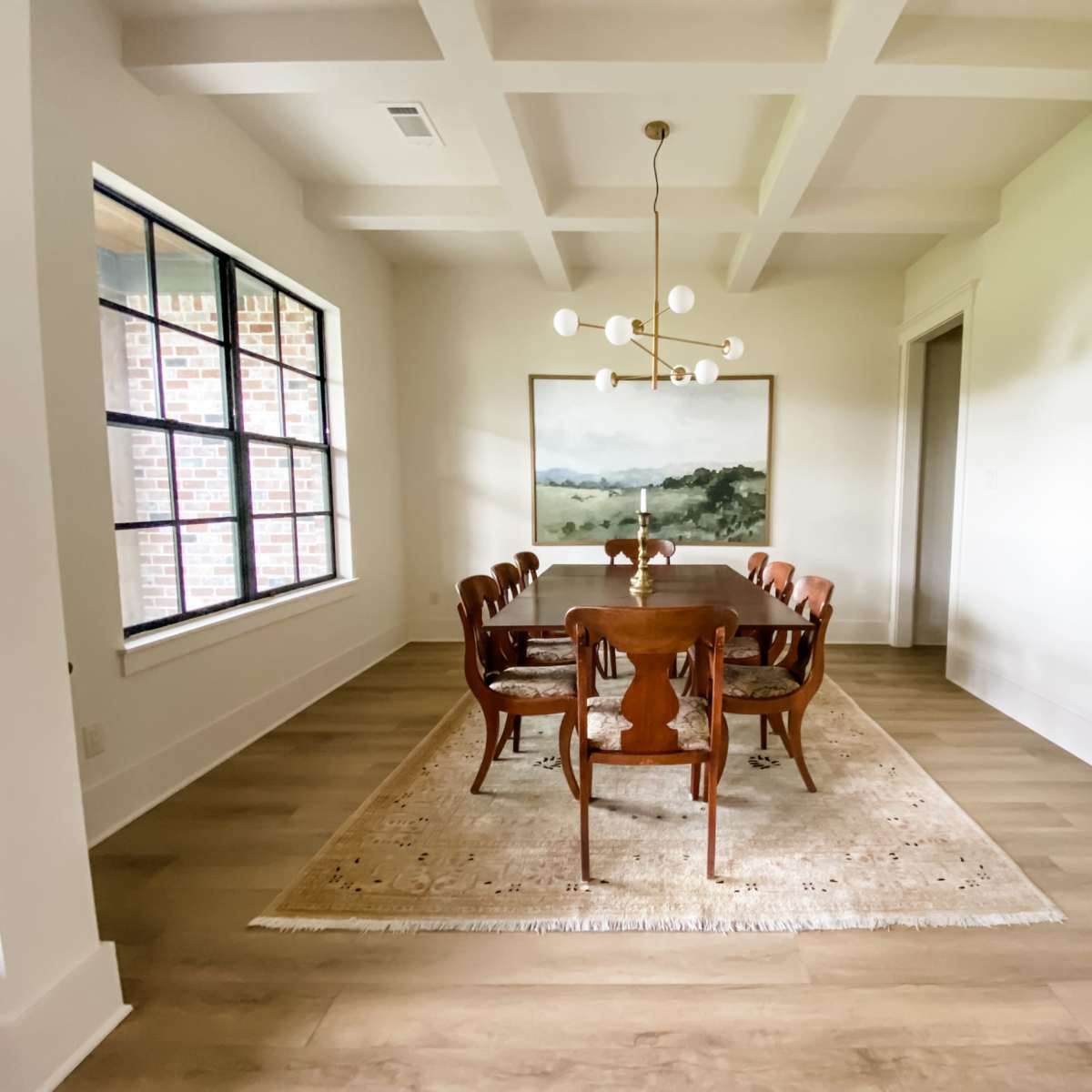 A dining room features a long wooden table surrounded by eight chairs, with a large window and a painting on the wall.