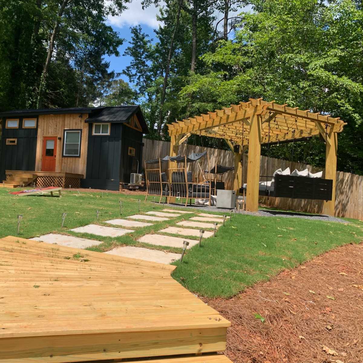 A small dark-colored cabin sits in a backyard with a wooden pergola, patio furniture, and a stone pathway leading through the grass.